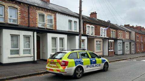 A marked police car parked on a residential street. The houses are terraced and mostly red brick. 