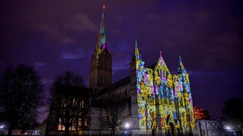 The front of a grand cathedral at night covered in a coloured light projection, the colours are yellow, blue and purple