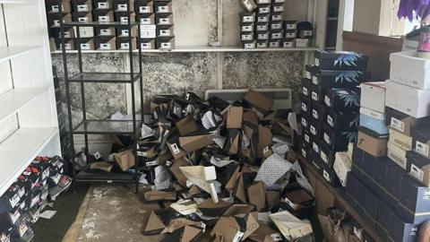 A room full of boxes of shoes that have fallen over and large black mould patches on the wall.