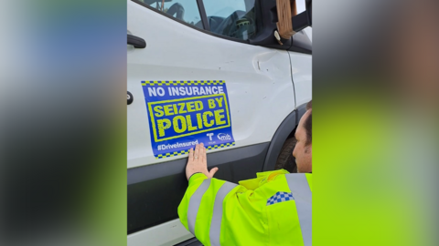 A man in a high-vis yellow jacket places a blue sticker on a white truck that reads "no insurance seized by police". 