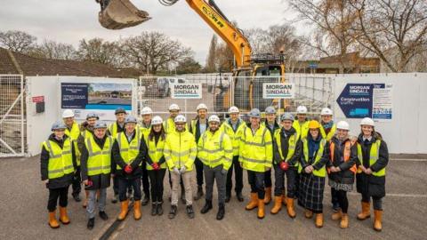 A group of people standing in front of a construction site wearing hi-vis jackets and hard helmets. They are all smiling at the camera.