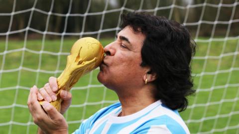 A man resembling Diego Maradona is stood in front of a football goal kissing a replica of the golden World Cup trophy. He is dressed in a blue and white football shirt, has thick black hair and wears an earing.