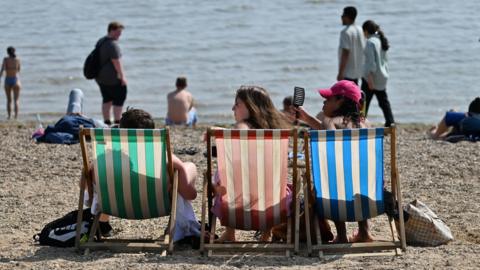 Beachgoers sit in deckchairs on the beach during the sunny hot weather.