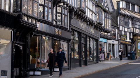 A street running up a hill with a series of black and white timbered buildings with shop windows running up the left hand side and a man and a woman walking down the hill