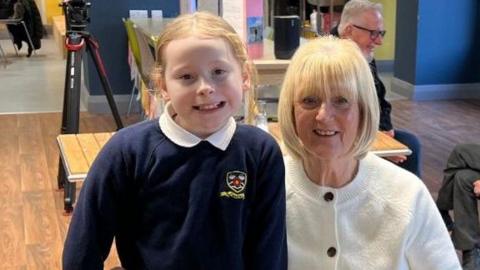 Lynne Aldersley, who is wearing a white cardigan, is sitting on a chair next to Skyla, who is standing. Skyla is wearing her school uniform which includes a dark blue jumper and white shirt. They are both smiling at the camera.