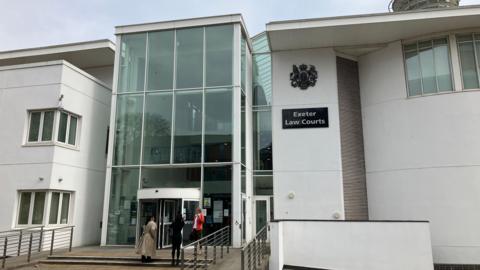 A white crown court building with a large glass entrance. A sign reads Exeter Law Courts. People stand on the steps.