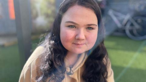 Charly Bisset - A young girl with long wavy dark hair sits outdoors in bright sunlight, wearing a light-coloured hoodie. A bicycle and greenery are visible in the background.