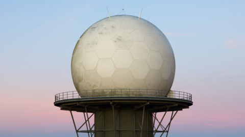A close up of a huge golf-ball like radar on top of a large platform with a fence around its perimeter. The dome is white and has hexagonal shapes on its outside, with small antenna on its top. Behind it is a pink and blue sky