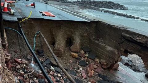 A view of the sink hole in a road with exposed rock and cables.