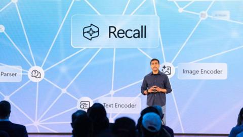 Microsoft executive Pavan Davuluri shown speaking on-stage about Recall at a Microsoft event, with the product's name featured on a display in the background.