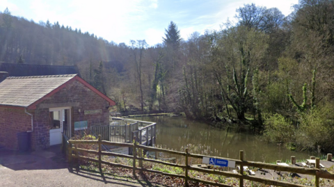 The Dean Heritage Centre overlooking a pond on a sunny day. It is surrounded by trees in a mini valley, and there is a wooden fence around the pond and a picnic area.