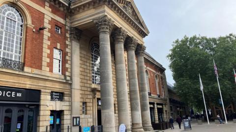 The outside of the Town Hall. Its grand entrance has four tall pillars holding a triangle-shaped structure. There are flags on poles in front of the building.