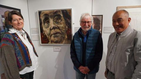 Three artists, one female two male, stood in front of a painting of a woman.