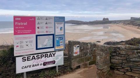 A picture of Fistral Beach in Newquay, Cornwall. There is a large sign providing lots of information and RNLI Lifeguard warnings. The beach is visible in the background with people walking on it. 