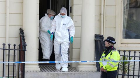 Forensic police officers leave a cream building, being guarded by a uniformed female police officer being a police tape cordon.