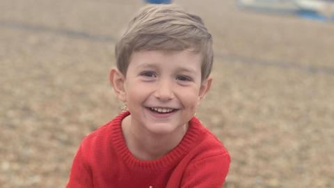 Benedict, a young boy with blond hair, is on a pebbly beach. He is wearing a red knitted top and is smiling to the camera.