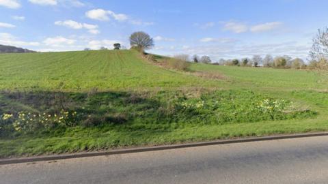 A grassy field seen from a road. It is a slope, with trees in the distance. The sky is blue.