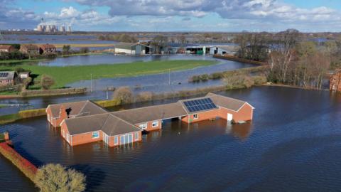 A village is almost entirely flooded by blue waters stretching almost to the horizon. In the foreground, a large red-brick bungalow property is surrounded by water. In the middle-distance are more homes and farm buildings, with patches of grass and trees. In the background, a large power station sits on the horizon under a blue sky.