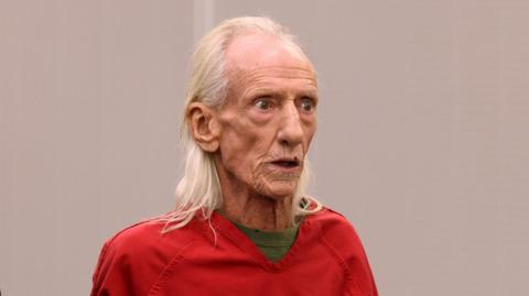 Joseph Czuba wears red overalls and a green shirt underneath. He has white, bedraggled hair.