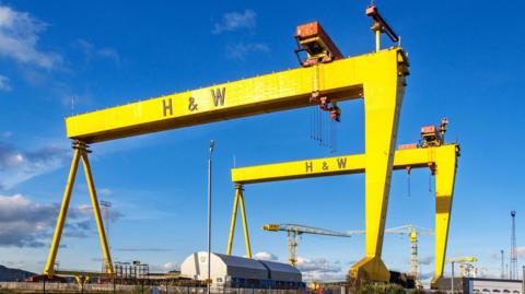 Two large yellow cranes with H & W written on them. Below them some smaller cranes and warehouses. Above them blue skies with a few clouds.