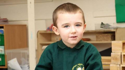 Lincoln Button is wearing a green school top.  He is in a schoolroom, surrounded by various bits of bric a brac.