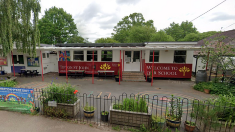 A google street view image of Tipton St John Primary School. The school building is half white half red. Over the red paint it red writing which said "Tipton St John" and "welcome to our school". 