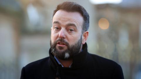 Headshot of Colum Eastwood speaking. He is wearing a black coat. the background is out of focus.