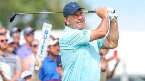 Sir Nick Faldo wearing a light blue t-shirt and a dark blue baseball cap holds a golf club over his shoulder