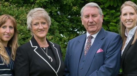 Aubrey Kirkham pictured in a blue suit with a pink pocket square stood next to wife Ann who wears a black suit and silver jewellery and their eldest daughter Leanne who is also smiling and wearing a blue and white striped shirt.