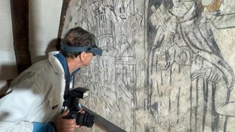 Mr Martindale is wearing high powered goggles or a type of glasses to look very close up at the painting while dressed in white overalls. He has a camera in his hands. The wall painting can be seen.