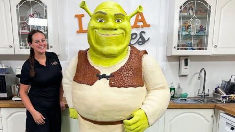 A woman with brown hair plaited over her shoulder, is wearing a black shirt and shorts and is smiling at the camera. She is standing next to a life-size cake replica of Shrek. Shrek looks as he does in the film series - with green skin and long ears and is wearing a long-sleeved white shirt and brown patterned vest.
© Lara Mason
