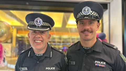 Chief Inspector Scott Collins with probationary constable Oliver Lamb. They are wearing uniforms and smiling. Scott Collins is a middle aged man and Oliver Lamb is slightly younger with a moustache. 