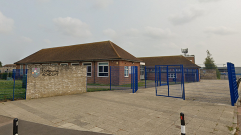 A yellow brick wall with the logo of The Eastbourne Academy in front of red brick, single-storey school buildings with sloped roofs. The school buildings are surrounded with a blue wire fence.