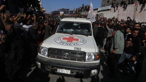 A red cross vehicle driving through a crowd of people. It is white with the red cross on the front.