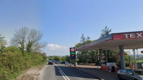 A rural two-lane road with hedgerows on each side, a Texaco garage sits on the right hand side of the image. 