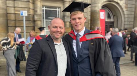 Two men stand beside each other outside an old stone building. The man on the left has very short hair and stubble wearing a black suit jacket and a grey zipped top. The man on the left is dressed in black and red graduation robes, graduation cap, suit jacket, a white shirt and dark grey tie.