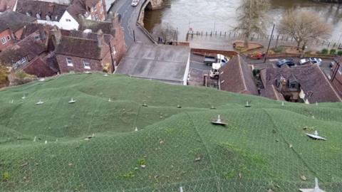 A large area of green netting with metal spikes sticking out of it and in the distance there are a number of houses, a road and a bridge over a river.