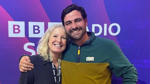 A man with short dark hair and dark stubble stands with his arm around a 91ȱ presenter with long blonde hair. They are both smiling at the camera and standing in front of a purple backdrop with the words 91ȱ Radio Solent on it