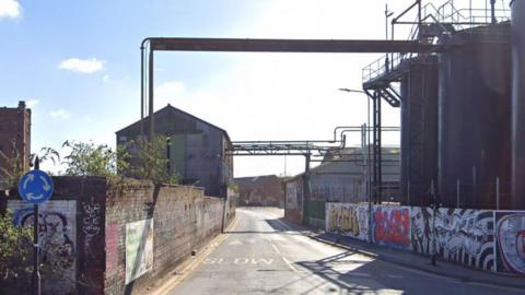 Two industrial buildings stand at either side of a road. Pipework stretching over the carriageway, between the two buildings, is visible. On the right side, two large black silos stand behind a wall covered in colourful graffiti.