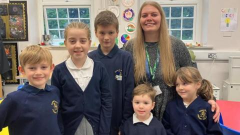 Pupils at Saxton Primary School breakfast club stand with breakfast club lead Jane. Pupils wear dark blue jumpers with white polo shirts.