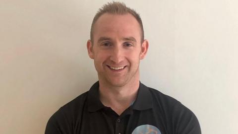 A man with short-cropped dark blonde hair smiles at the camera. He is wearing a black polo neck shirt.