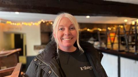 Angela McCafferty wearing a black t-shirt with a black coat over the top of it. She is smiling at the camera and is stood inside of her pub. There are brown bar stools on top of the bar and yellow lights hung across the ceiling. 