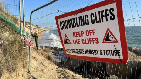 A cliff edge is blocked off by a metal fence. A sign on the fence in bold letters reads: "Crumbling cliffs. Keep off the cliffs. Help preserve Hemsby's future."