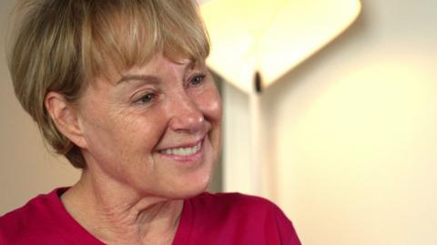 Sally Dynevor sits smiling at someone off camera. She is wearing a pink t-shirt. 