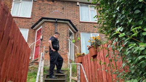 A police officer stands on a flight of stairs upto a house looking down. Behind him are two terraced houses, and either side of the steps are some white rails and then orange fences 