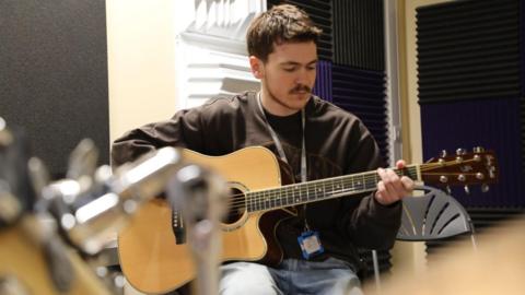 Teenage boy playing an acoustic guitar. He is wearing a black jumper and a lanyard around his neck and has a moustache and brown hair.