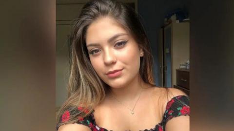 A young woman looks into the camera. She is wearing a black and red top and has long brown hair over her shoulders and brown eyes. 