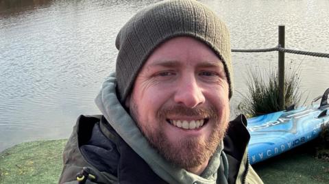 A man with a green beanie hat, a beard and a green coat, smiles as he takes a selfie. A lake is behind him.