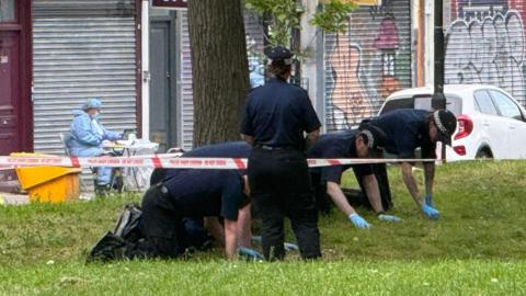 Police officers hunched on the ground with blue plastic gloves going through the grass for evidence. You can see police tape in the front and in the back is a forensic officer. 
