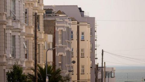 A row of light-coloured terraced buildings on a clear day. The sea can be seen in the background.